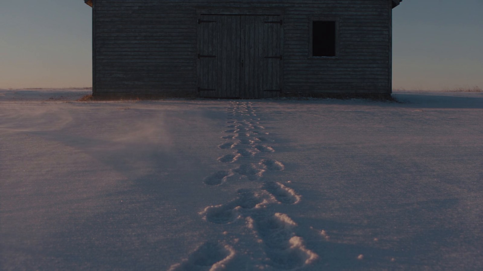 A single set of tire tracks in light snow leading to a shuttered clapboard cabin at dusk.